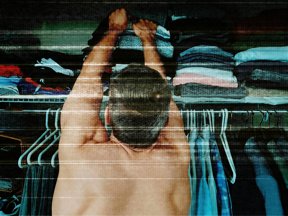 Person standing in front of a closet, reaching up to a shelf with various folded clothes