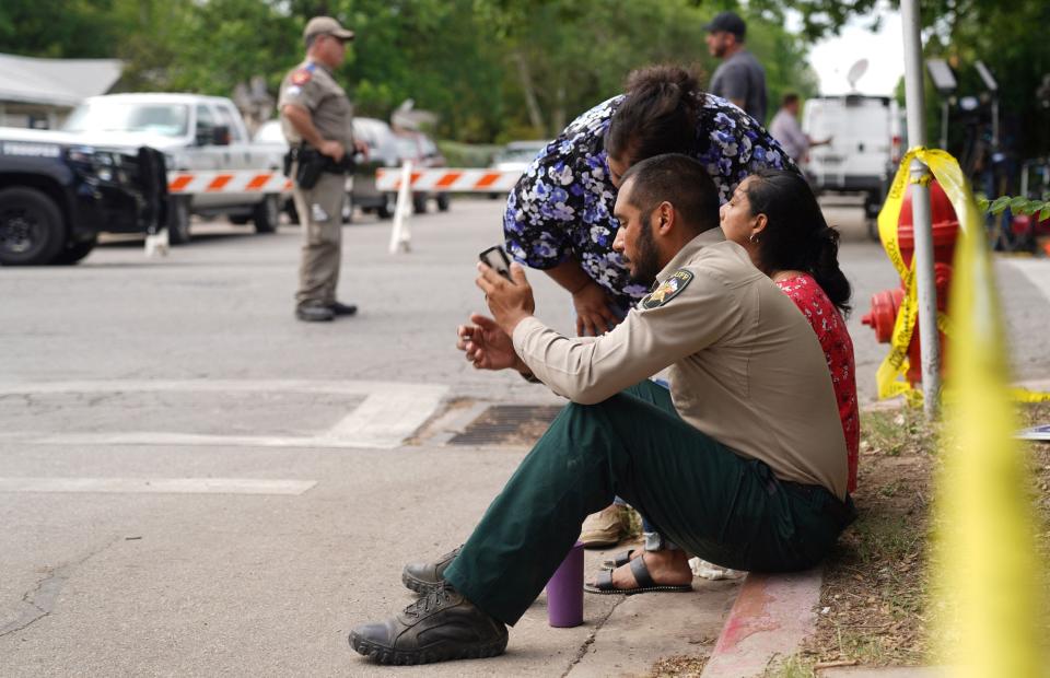 A sheriff checks his phone as he sits on the sidewalk with two women outside Robb Elementary School as state troopers monitor the area in Uvalde, Texas, May 24, 2022.