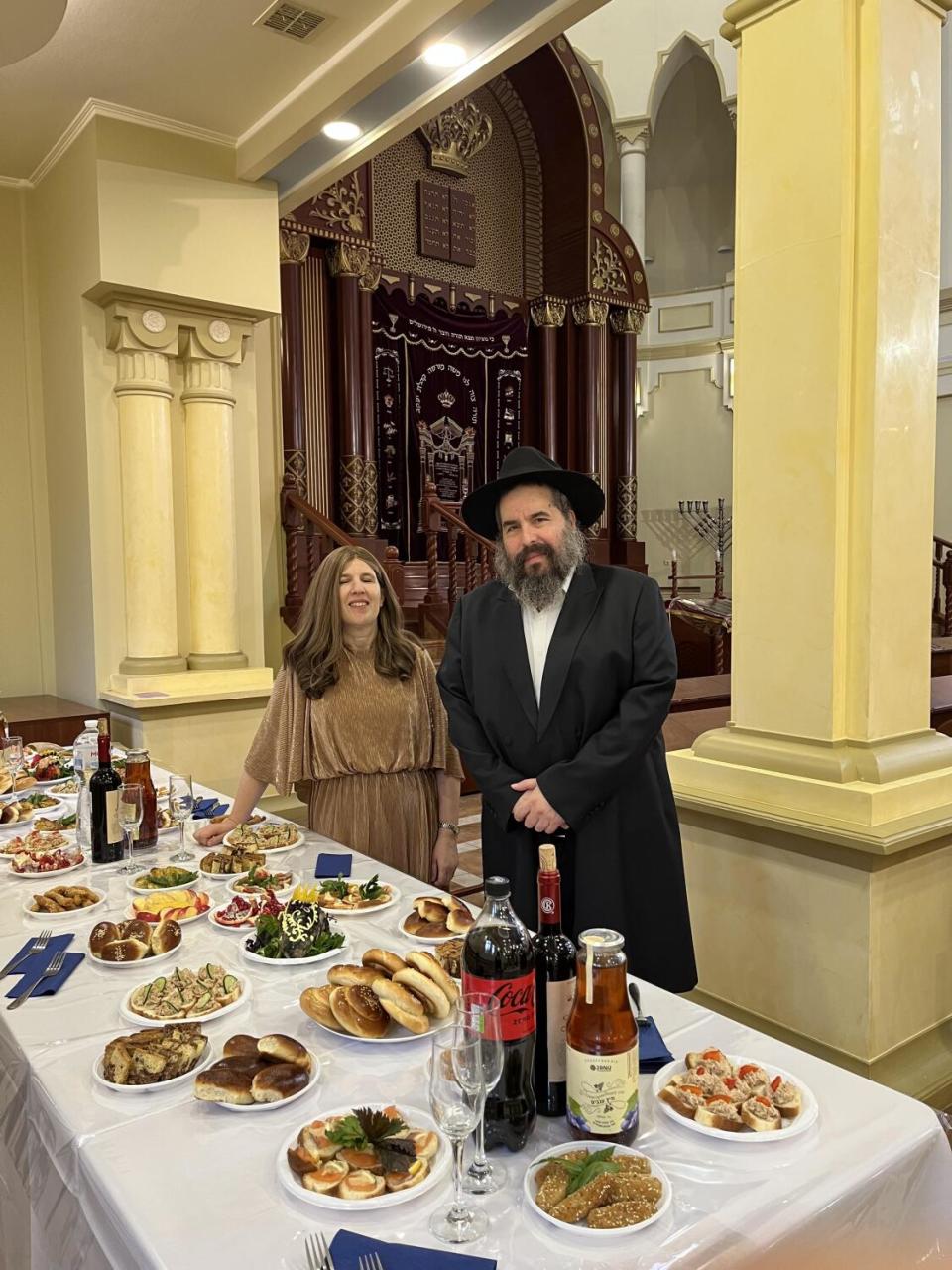 A woman in a brown dress and a bearded man in dark clothing and hat stand before a table laden with plates of food