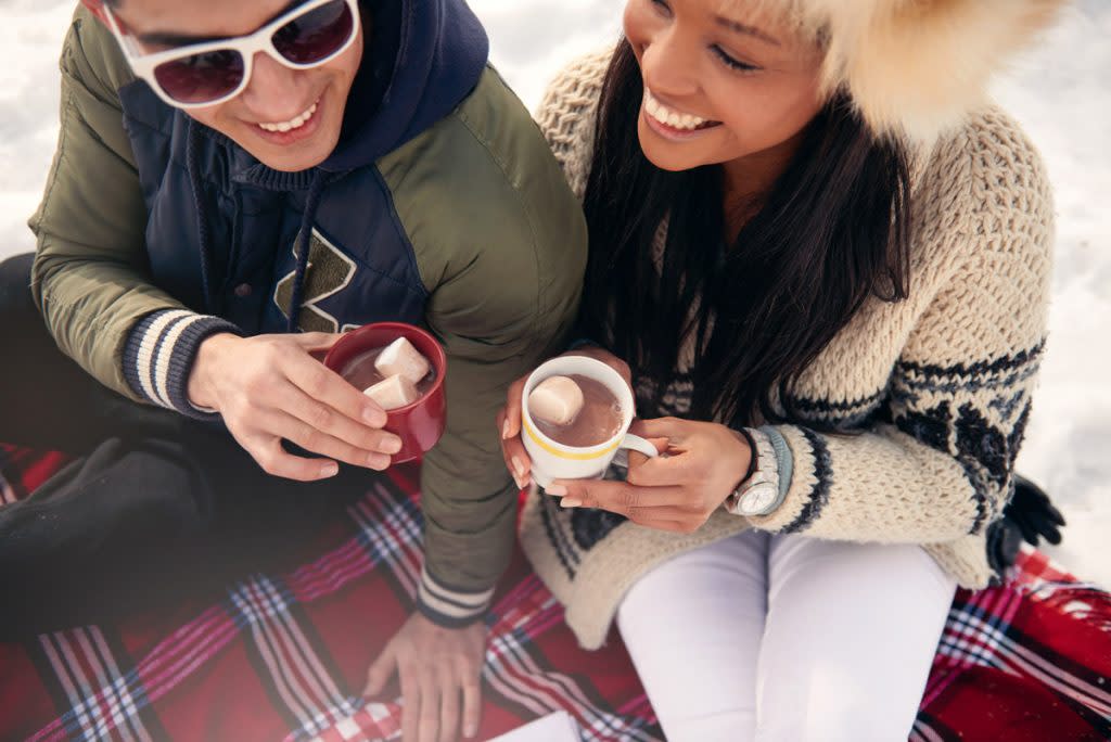 A couple sit on a blanket in the snow while sipping on hot chocolate. 