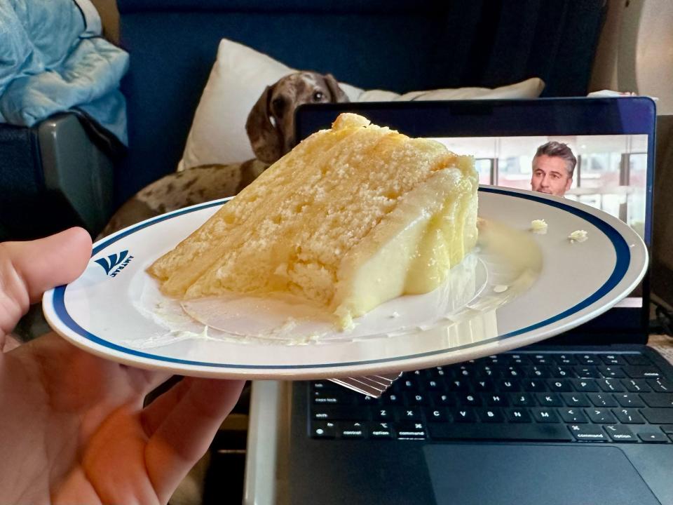 A hand holding a plate of lemon-custard cake in front of a laptop. Sean's dog Oliver can be seen lounging on a pillow behind his laptop.