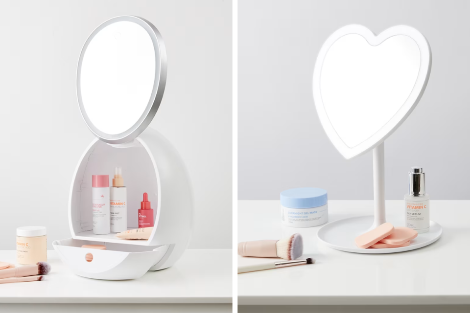 L: Oval makeup mirror with storage from Kmart containing beauty products on a grey background. R: Heart shaped LED white makeup mirror from Kmart with beauty products on the table against a grey background