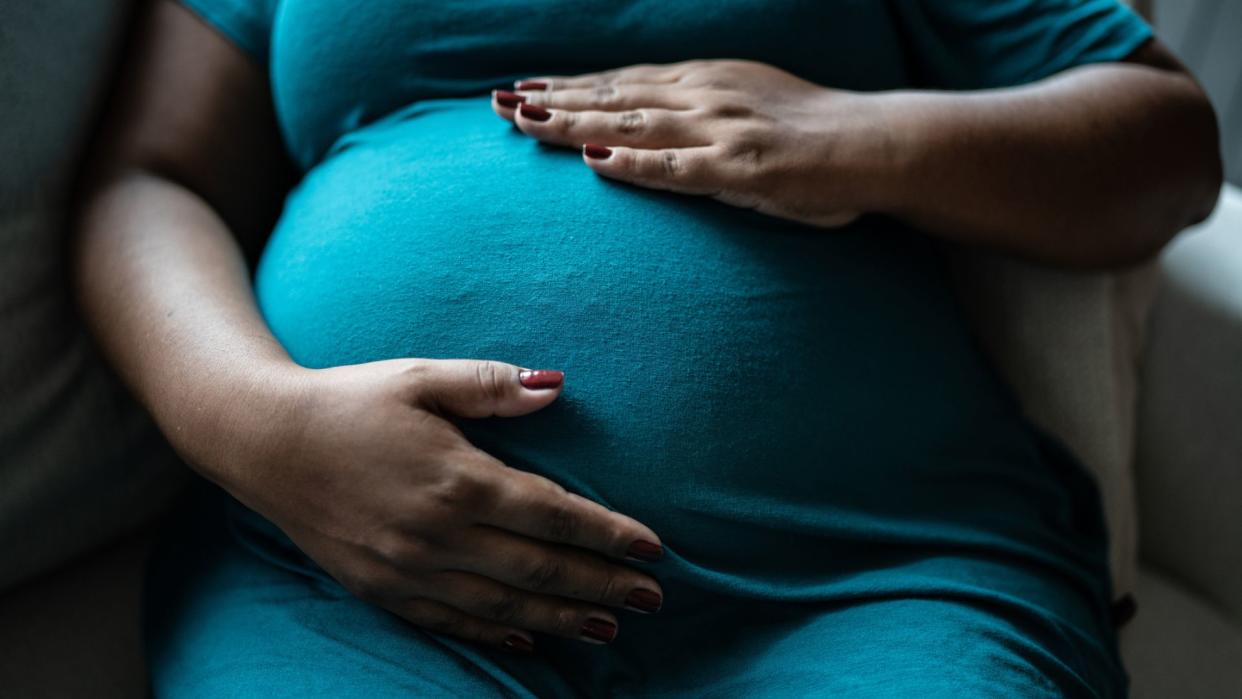  close up of a pregnant Black woman's belly. She wears a teal dress and has her hands gently placed on top and bottom of her stomach 