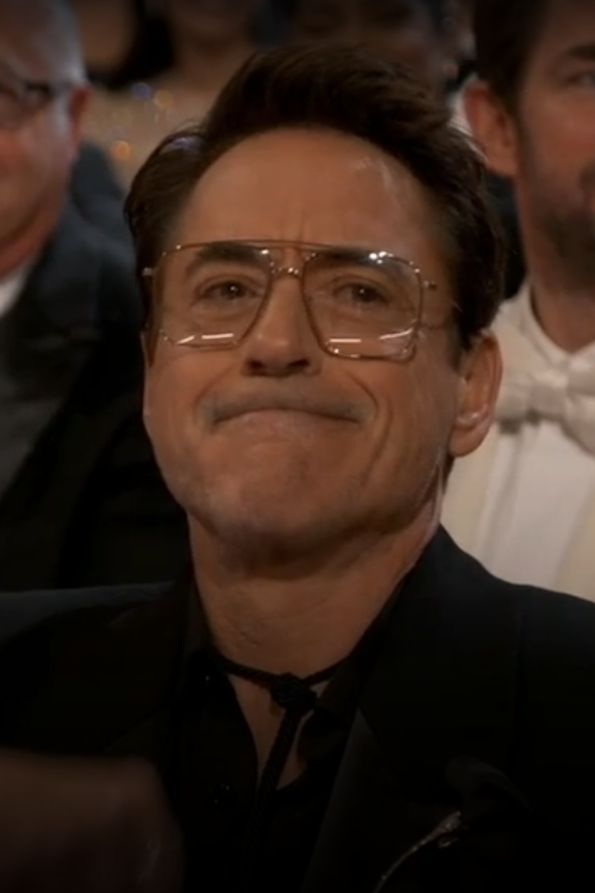 A smiling man in glasses and a tuxedo seated at an event, flanked by attendees in formal wear