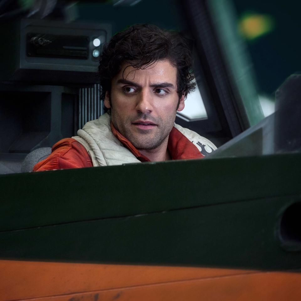 Oscar Isaac, dressed as his character Poe Dameron in a futuristic outfit, is seated inside a spacecraft cockpit