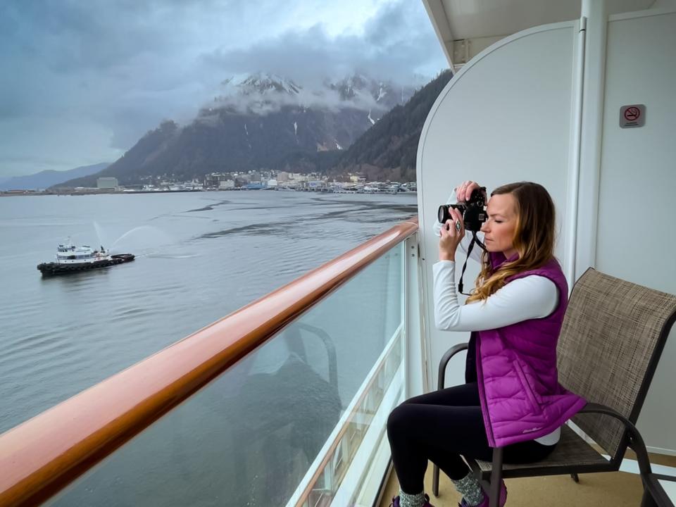 Emily, wearing a purple puffer vest, a white long-sleeve shirt, black pants, and purple sneakers, takes a photo on the balcony of an Alaskan cruise.