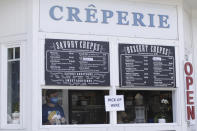 In this Thursday, May 28, 2020, photo, Yuri Vidal, co-owner of Crepe Neptune, wearing a face mask due to the coronavirus as he holds a customer's order at the take-out window of the creperie in Cannon Beach, Ore. With summer looming, Cannon Beach and thousands of other small, tourist-dependent towns nationwide are struggling to balance fears of contagion with their economic survival in what could be a make-or-break summer. (AP Photo/Gillian Flaccus)