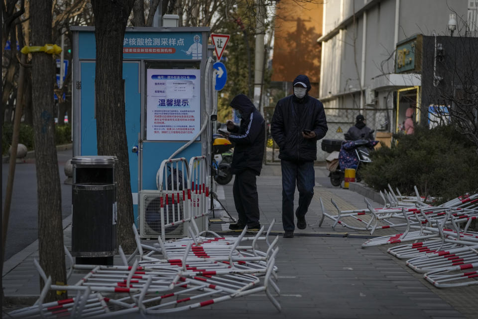 A masked man walks by a resident who registers to get his COVID-19 throat swab near barricades laying around a coronavirus testing site in Beijing, Wednesday, Dec. 14, 2022. China's National Health Commission scaled down its daily COVID-19 report starting Wednesday in response to a sharp decline in PCR testing since the government eased antivirus measures after daily cases hit record highs. (AP Photo/Andy Wong)