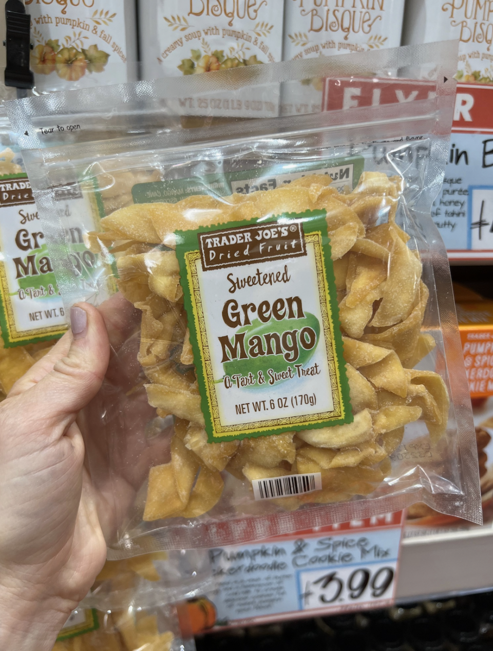 A hand holding a bag of Trader Joe's Sweetened Green Mango slices in a grocery store. Pumpkin products are visible in the background