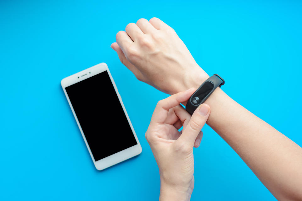 Close-up shot of one hand adjusting a fitness tracker worn on the other wrist. Below the hands, a smartphone lies on a sky-blue surface.