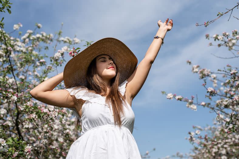 young caucasian woman with her eyes shut wearing a white summer dress next to a flowered apple tree to demonstrate the cottagecore trend