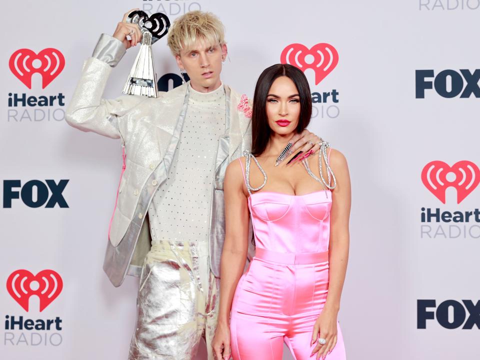Rapper Machine Gun Kelly wears a white and silver suit and holds an iHeart Radio Music award while posing with his hand on girlfriend Megan Fox's neck, who is wearing a pink satin jumpsuit and staring at the camera.