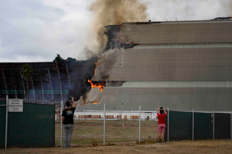 Two spectators take pictures as a historic blimp hangar burns in Tustin, Calif., Tuesday, Nov. 7, 2023. A fire destroyed a massive World War II-era wooden hangar that was built to house military blimps based in Southern California. (AP Photo/Jae C. Hong)