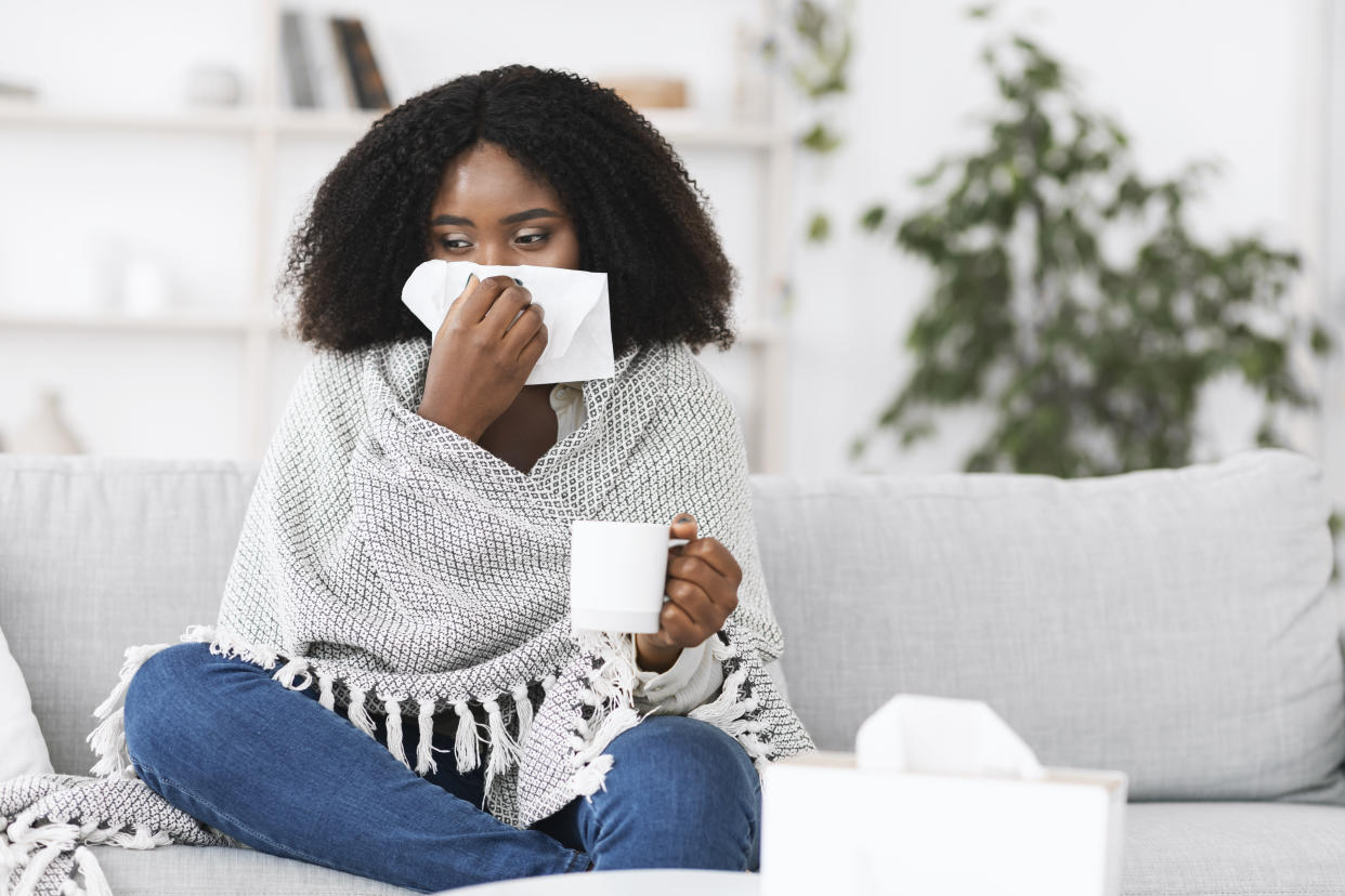 Fever, Cold, Flu Symptoms And Folk Medicine. Sick ill black woman covered in blanket blowing her running nose in a tissue sitting on sofa, drinking hot tea, holding cup, copy space. Taking medication