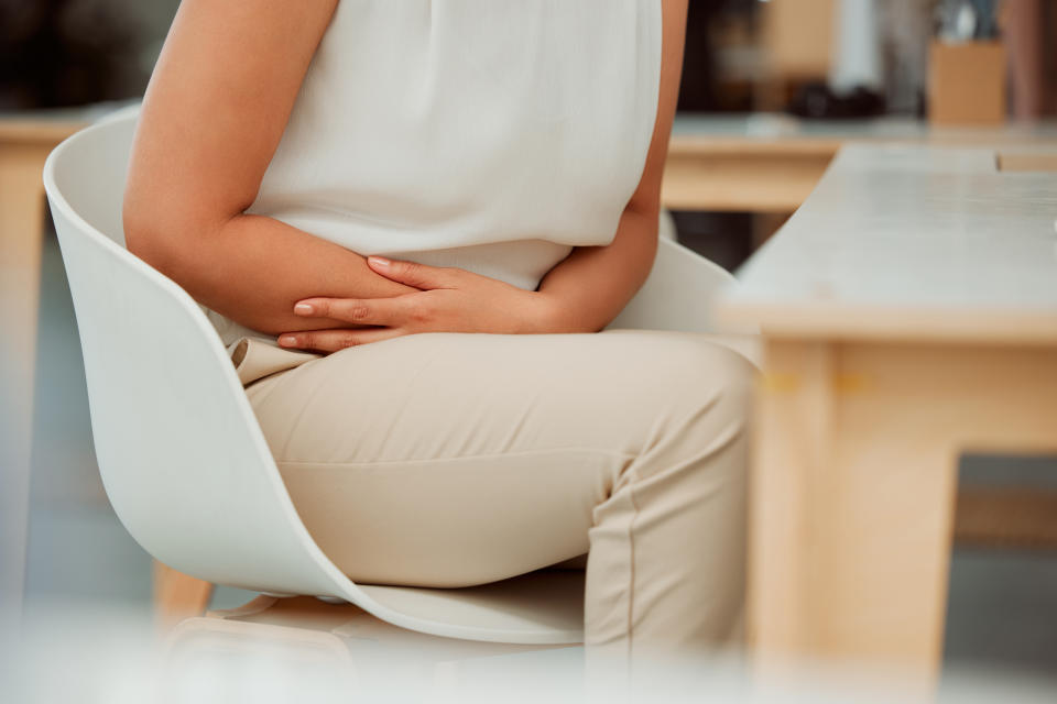 fertility  Closeup of one mixed race business woman holding sore tummy while feeling ill with menstrual stomach cramps and belly ache in an office. Hungry employee on period getting sick, bloated and uncomfortable with digestive pain caused by stress and anxiety