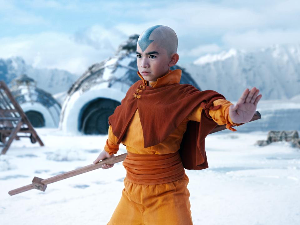 aang in the live action netflix avatar the last airbender. a young boy wearing red and yellow loose clothing, holding a wooden staff, and with a blue arrow tattooed on his forehead