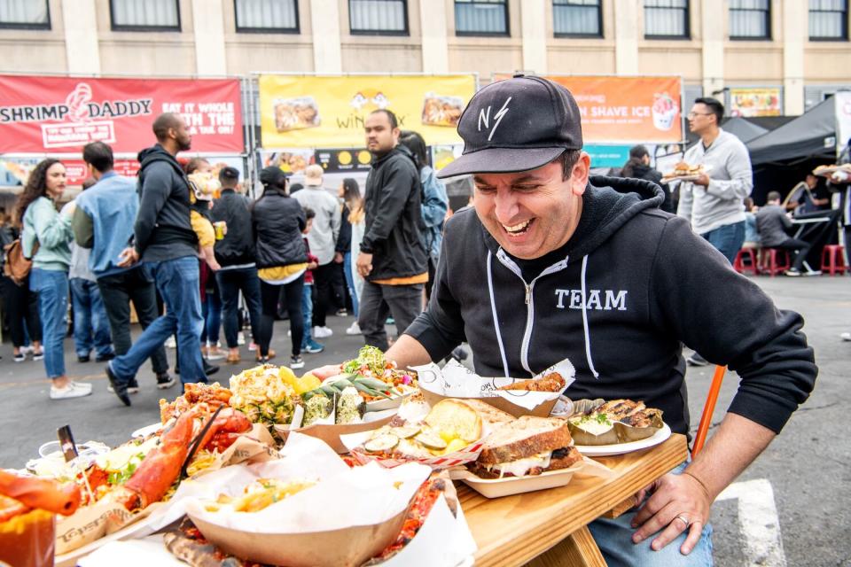 A smiling man in a black baseball cap sits in front of a table full of food.