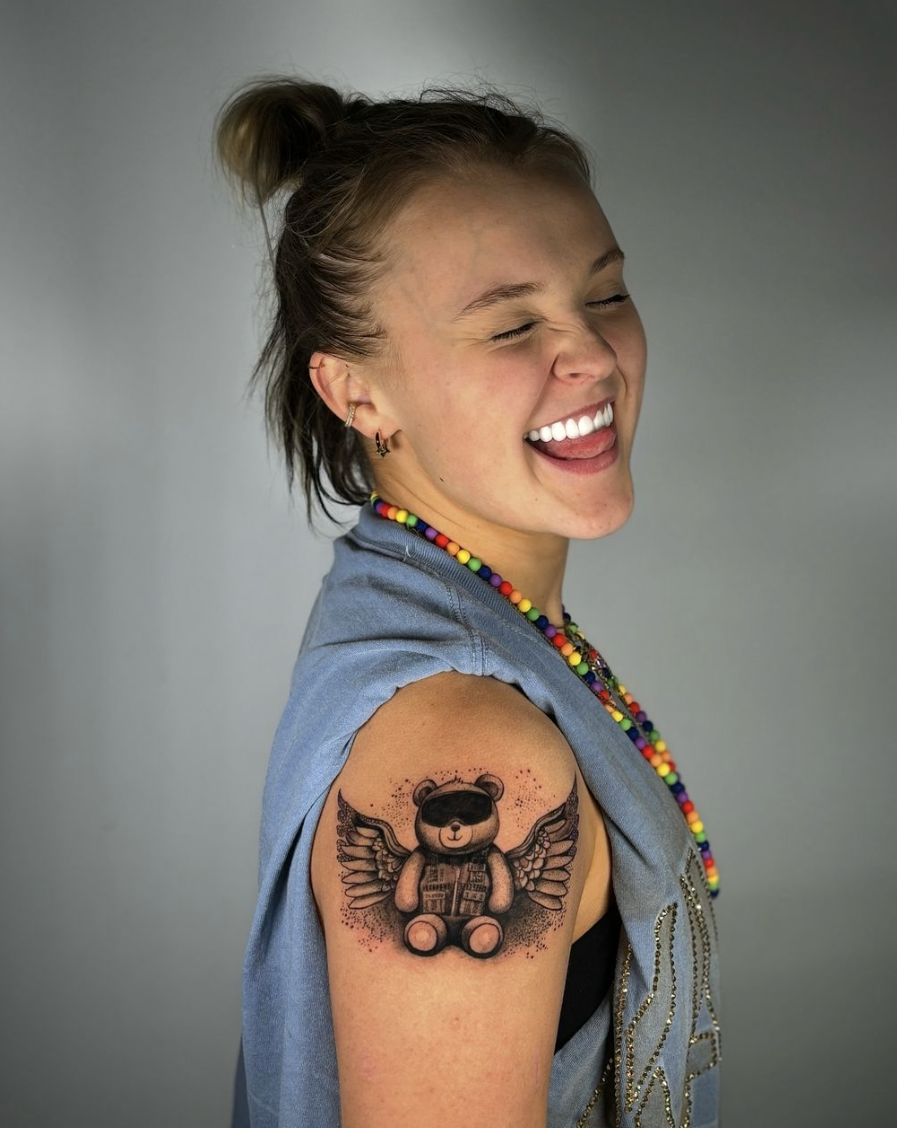 JoJo Siwa smiling with eyes closed, showing a tattoo of a teddy bear with wings on her upper arm. She is wearing a sleeveless top and colorful bead necklace