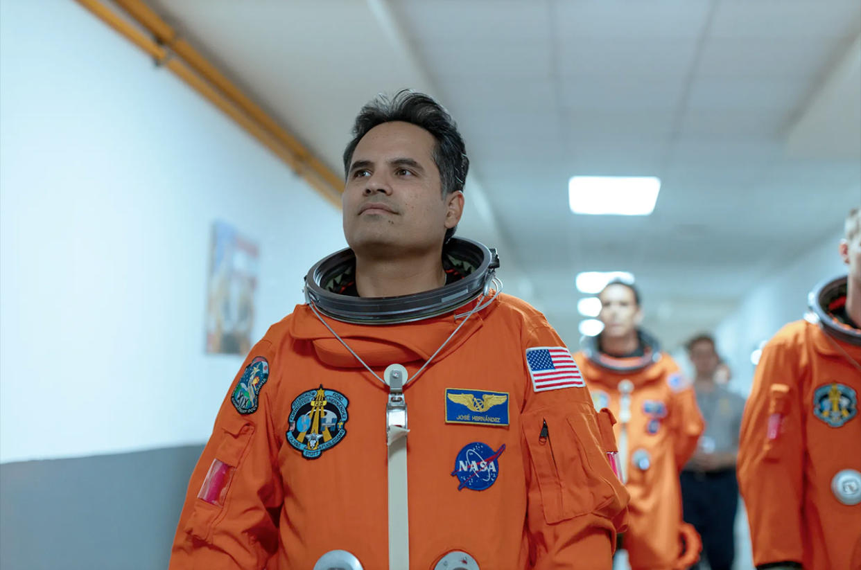  three astronauts in orange flight suits walk down a white hallway. 