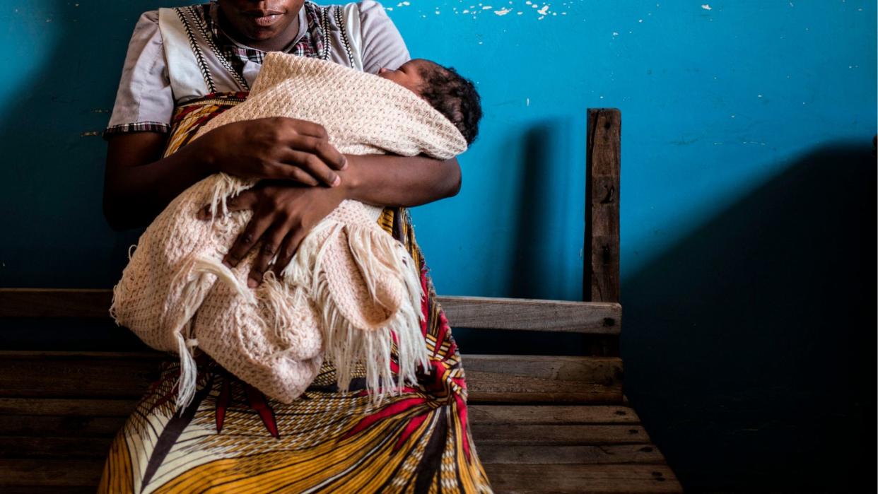  A woman sits on a bench holding a baby wrapped in a blanket, whose face is turned from the camera. 