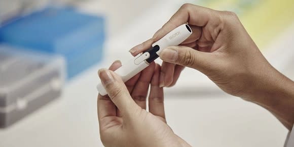 Two hands holding an iQOS heated tobacco unit device in front of an out-of-focus table.