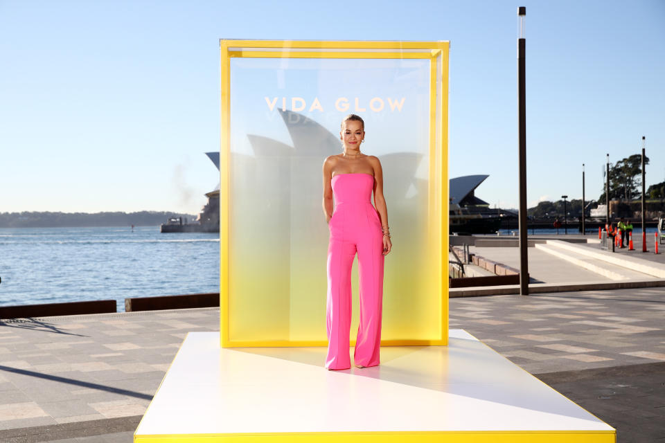 Rita Ora in pink strapless jumpsuit stands on an outside yellow and white stage before a yellow screen advertising Vida Glow, at Sydney Harbour on May 17, 2021 in Sydney, Australia. 