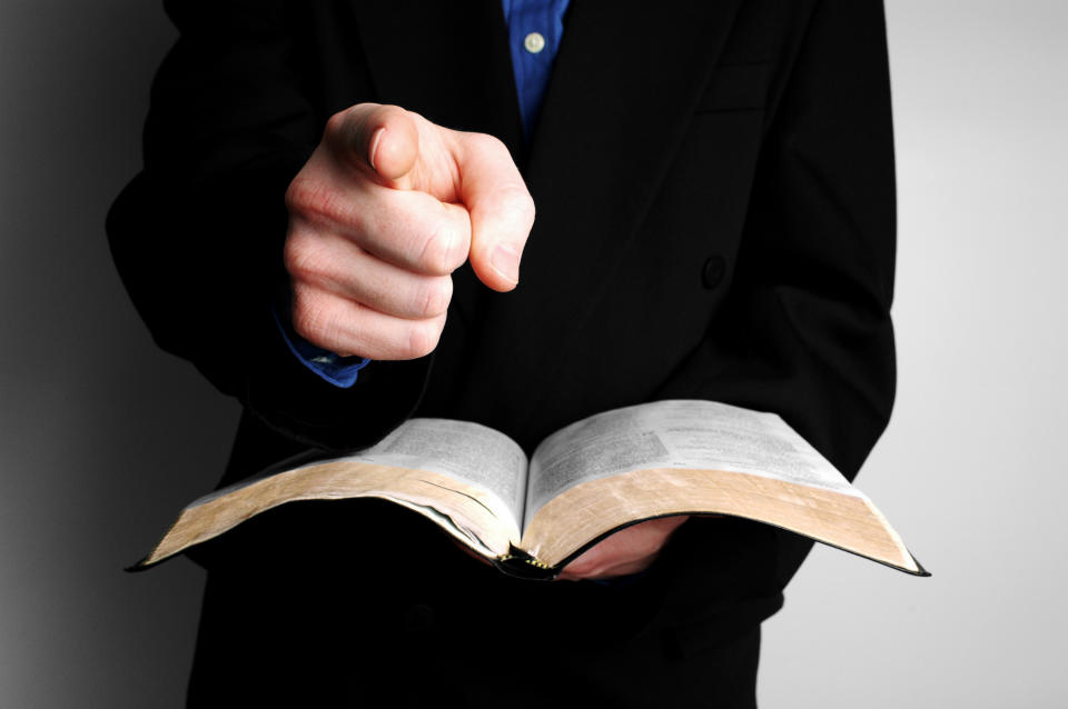 A person in a black suit points directly at the camera while holding an open book. The focus is on the hand and book