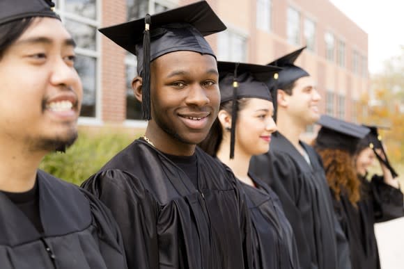 Row of college graduates in black gowns smiling on a hazy day in front of a building.