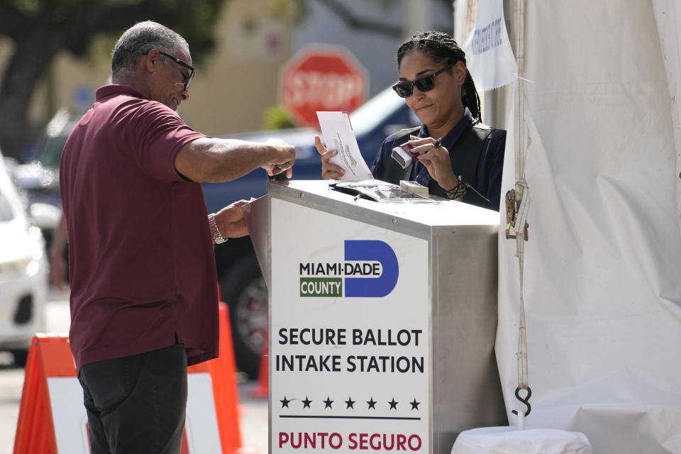 Employees process vote-by-mail ballots at a desk that says: Miami-Dade County Secure Ballot Intake Station Punto Seguro.