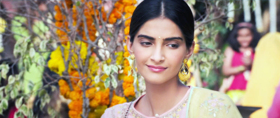 Sonam Kapoor wearing traditional attire with gold earrings and a bindi, stands with flowers and foliage in the background