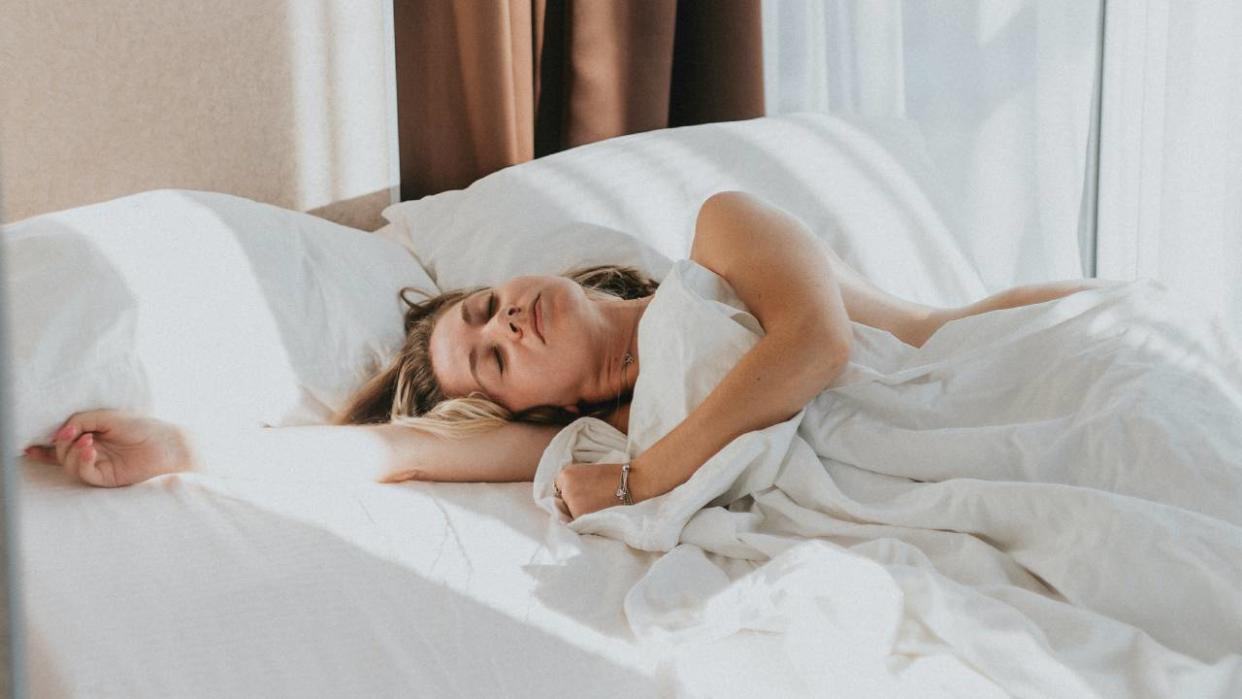  A woman asleep while lying in bed with white sheets, pillows and duvet. 