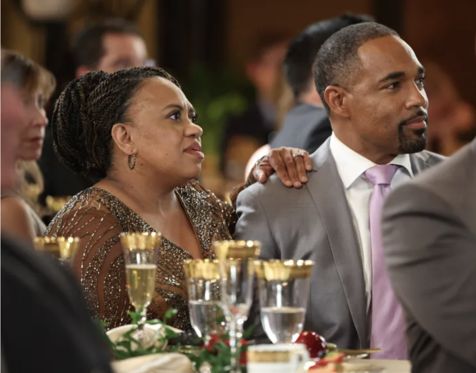 Two people at an event table, woman wearing sequined gown, man in suit, both looking to the side