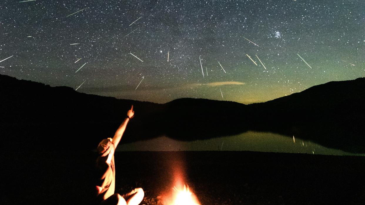   A figure sitting next to a campfire points up at a starry sky, streaked with shooting stars. 