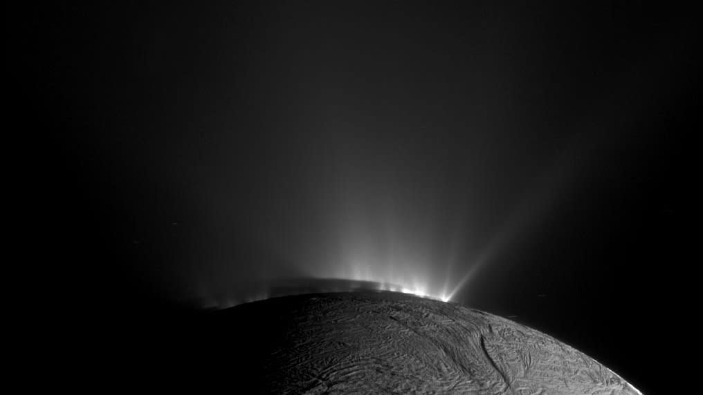  The top of a planetary body rises in black and white from the bottom. light illuminates streaks of geysers spewing from the surface near the top. 