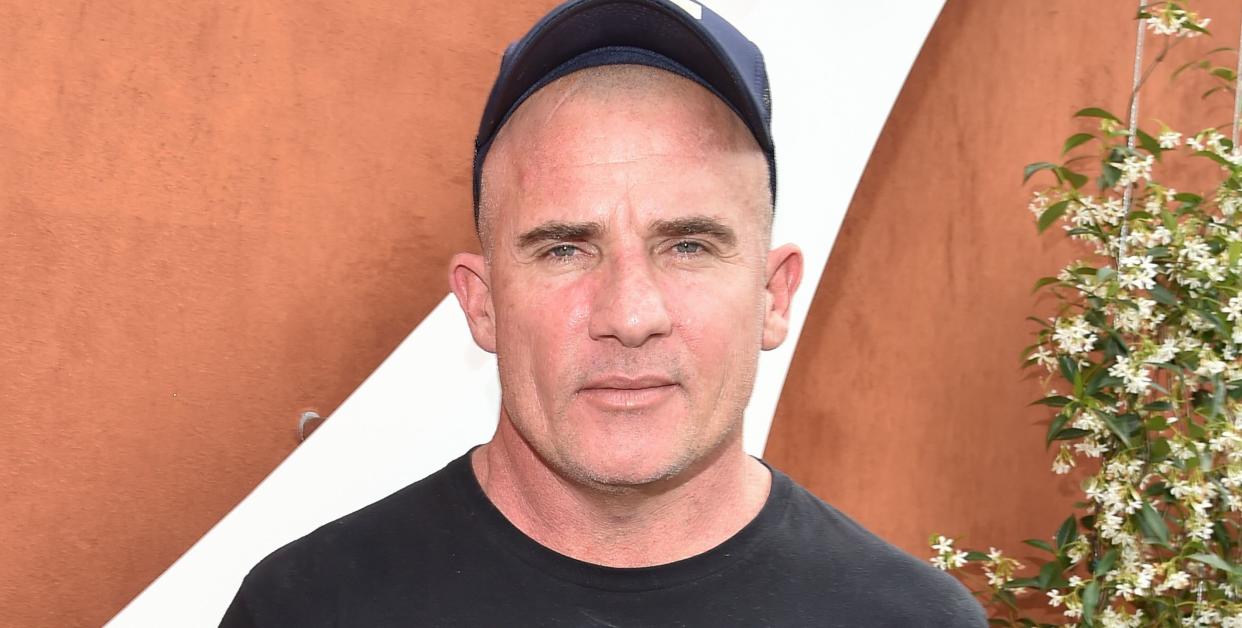 dominic purcell, a man stands looking at the camera with a neutral facial expression, he wears black jeans and tshirt with a black cap on his bald head