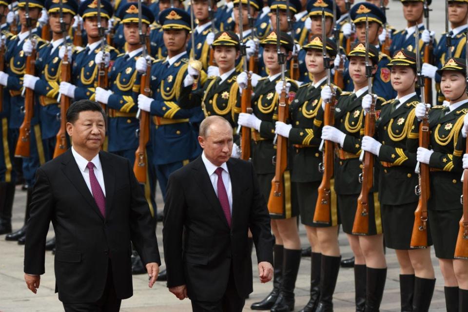 Xi Jinping and Vladimir Putin in suits walking in front of lines of troops in uniform holding rifles.