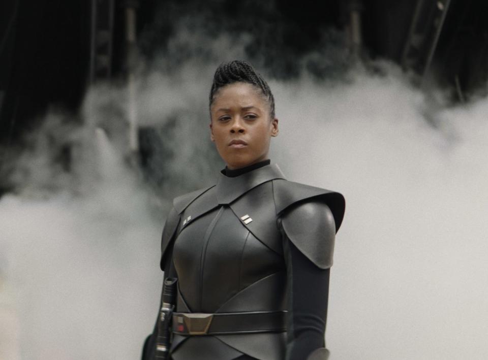 Moses Ingram stands in front of a smoky background, wearing a futuristic black armored outfit with angular shoulder pads