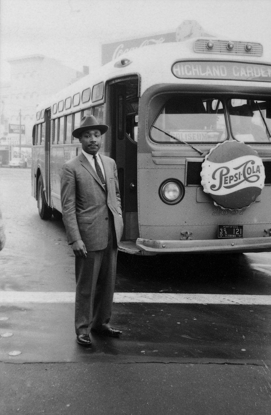 martin luther king jr stands outside in a suit and hat, behind him is a city bus with a pepsi cola ad on the front