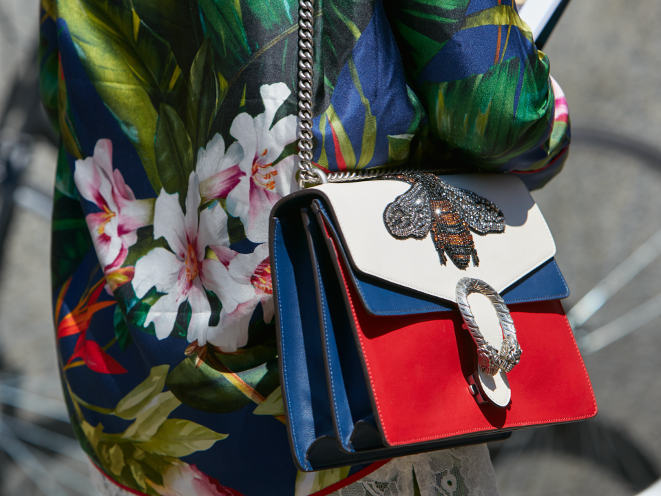 Frau, die eine Straße entlanggeht, eine Jacke mit tropischen Motiven trägt und eine rot-weiß-blaue Handtasche mit big embroidered and metal accents