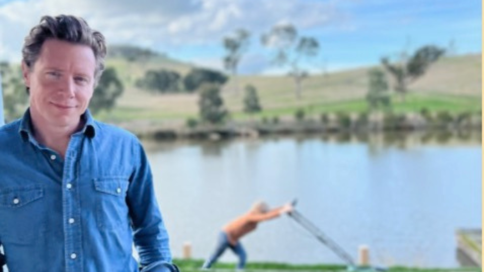 Barefoot Investor Scott Pape looks into the camera while a kid mows the grass in the background.