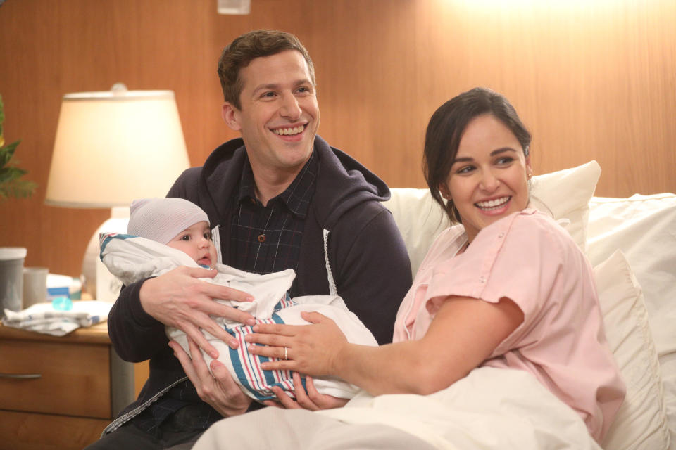Jake Peralta and Amy Santiago from Brooklyn Nine-Nine smiling, holding their baby in a crib