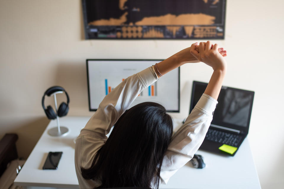 A woman stretching at her desk in front of a computer and laptop