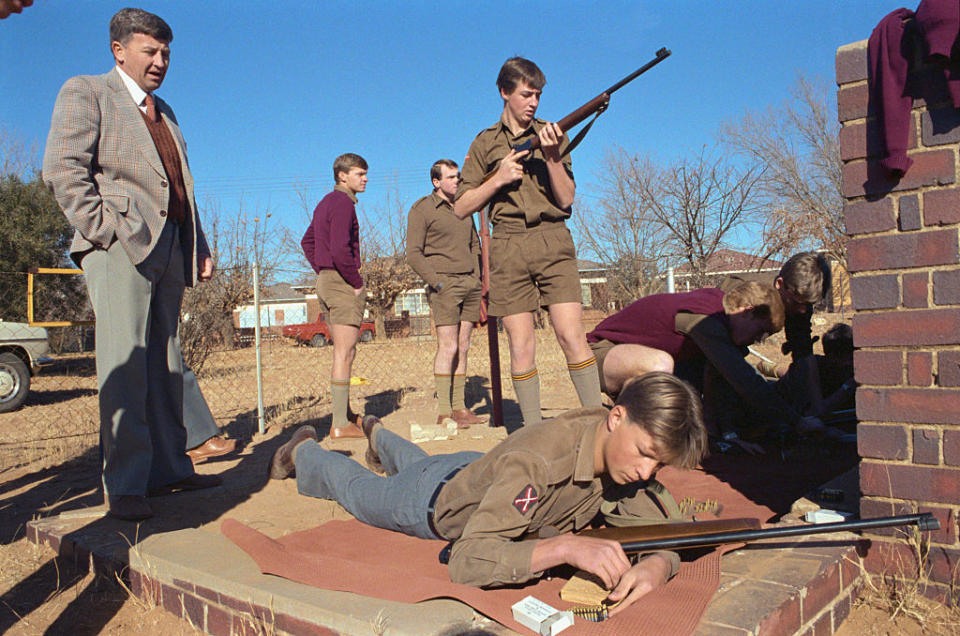 A group of boys in uniform practice shooting under adult supervision at an outdoor range. One boy aims a rifle while another lies on the ground