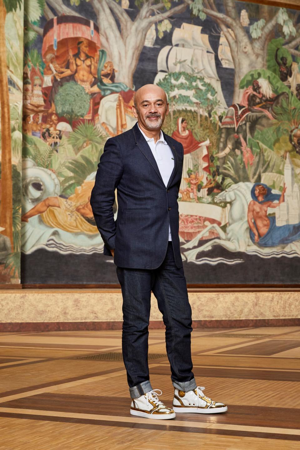 Designer Christian Louboutin dressed in blazer, jeans and sneakers, standing in front of a wall size piece of art.