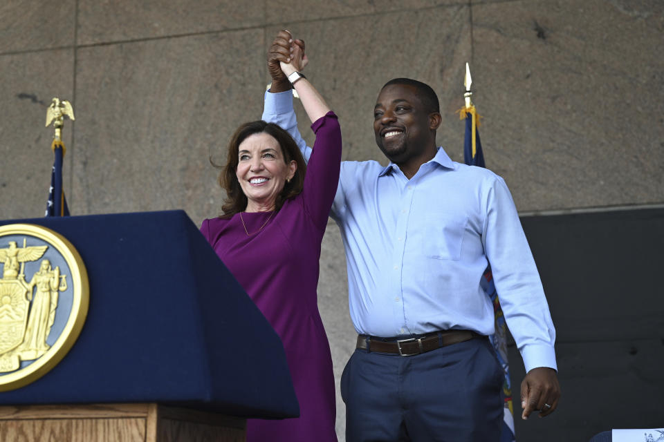 Kathy Hochul and Brian Benjamin clasp each other's hands over their heads behind a podium with the New York State seal.