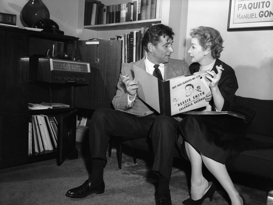 Leonard Bernstein and Felicia Montealegre sit on a couch, both holding cigarettes and a record album, in this 1956 black and white photo.