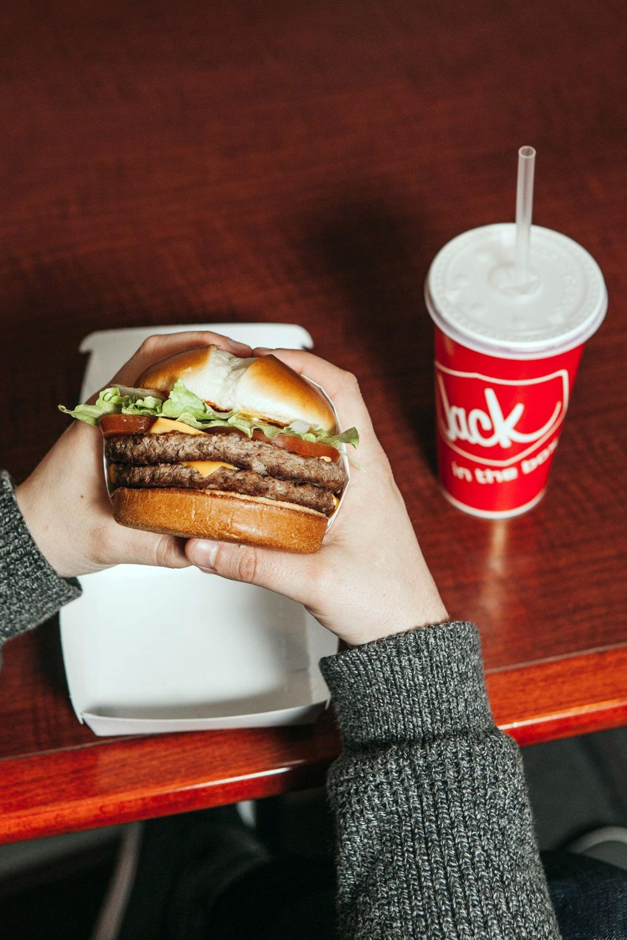 hands holding jack in the box cheeseburger, drink in background