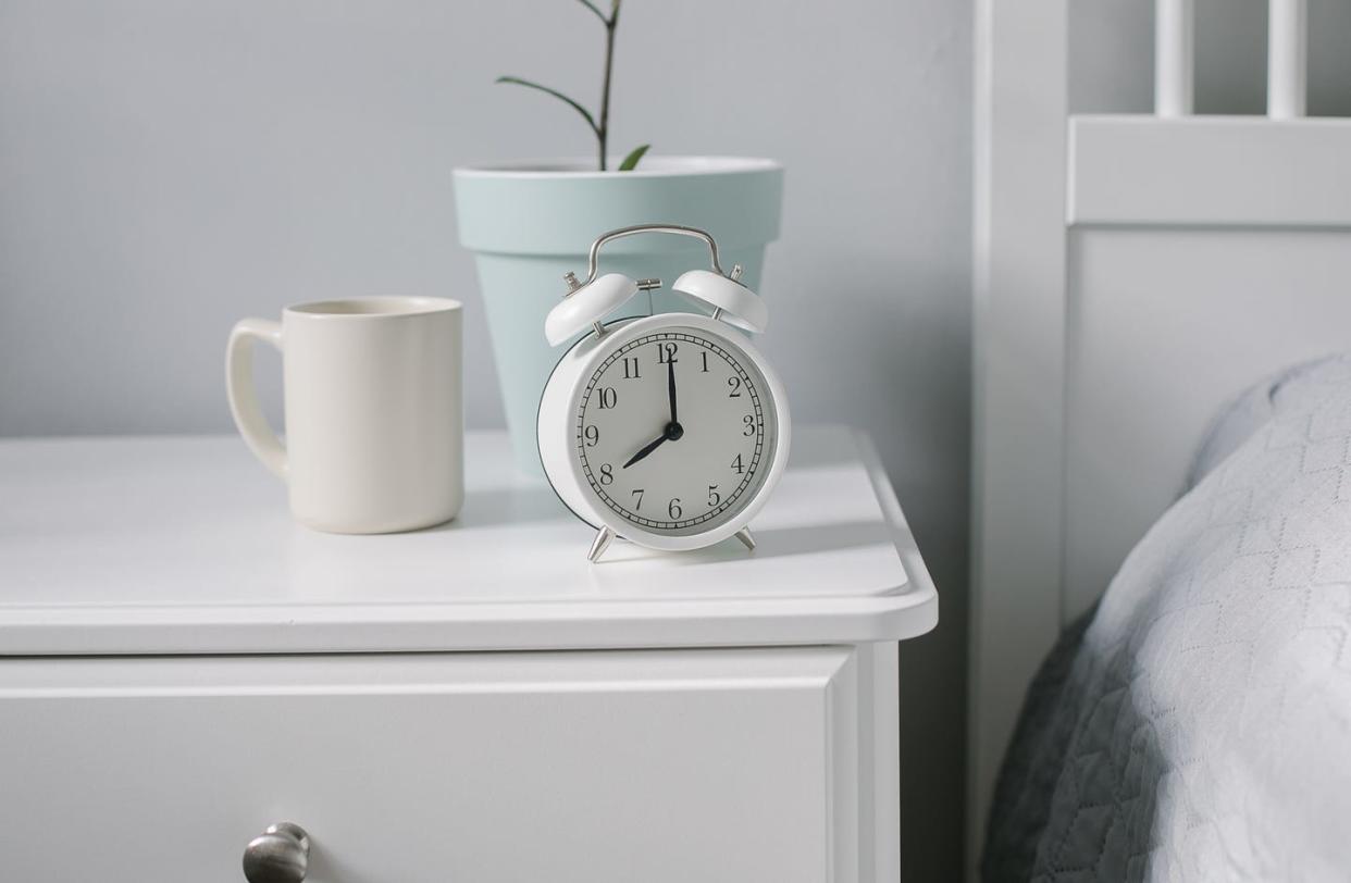 white alarm clock, beige cup with coffee, tea, house plant on bedsite in bedroom morning concept