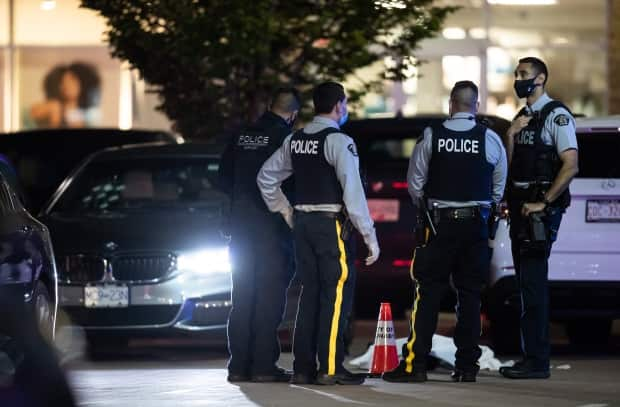 RCMP officers stand near a body covered with a tarp as bullet holes are seen in the windshield of a vehicle in the parking lot of the Market Crossing shopping complex in Burnaby, B.C., Thursday night. (Darryl Dyck/The Canadian Press - image credit)