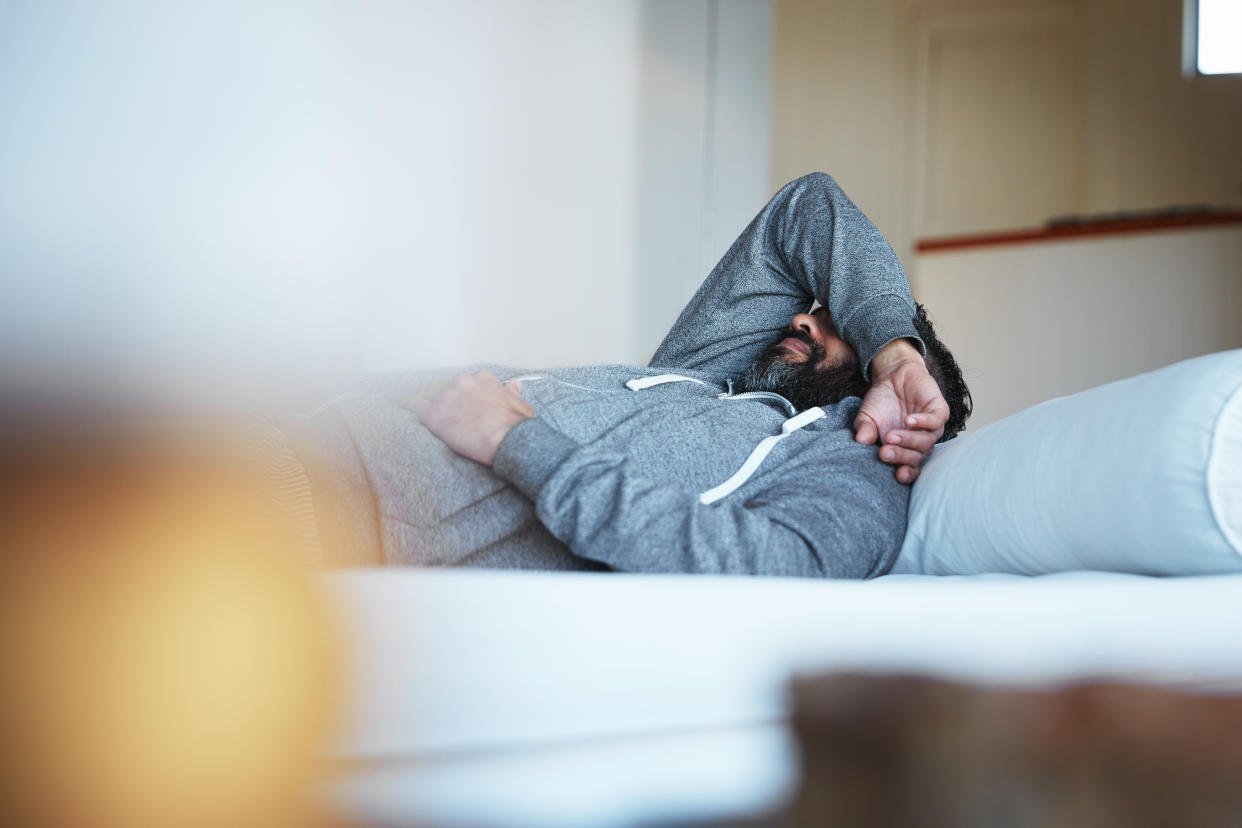 A man wearing a grey hoodie lays on his bed with an arm over his face